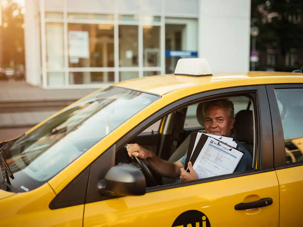 Taxichauffeur in gele cab voor Nederlands arbeidsbureau, met officiële documenten op de passagiersstoel en hand aan het stuur.